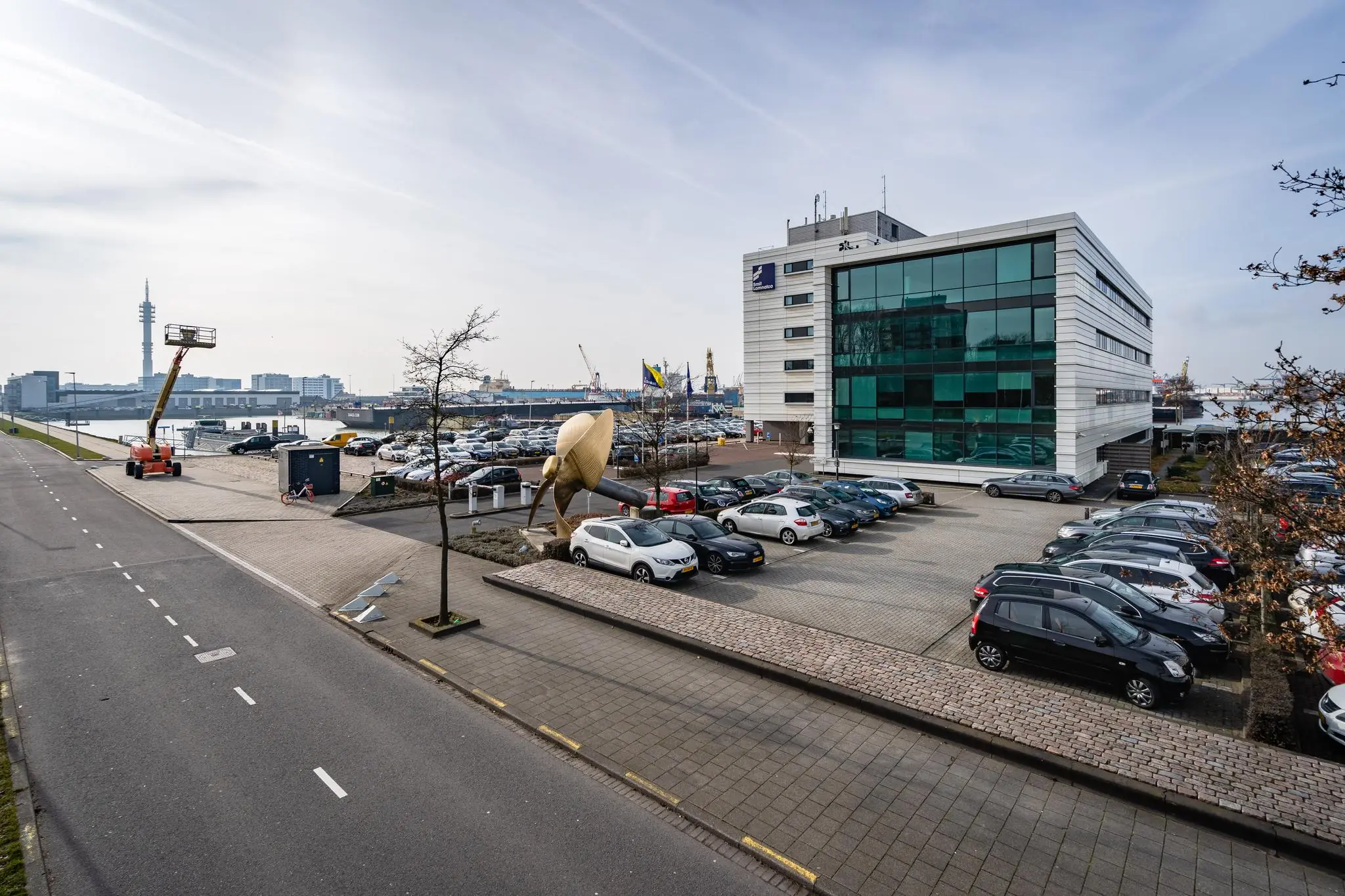 Kantoorgebouw met glazen gevel en parkeerterrein aan de Waalhaven in Rotterdam, met op de achtergrond havenkranen en schepen.